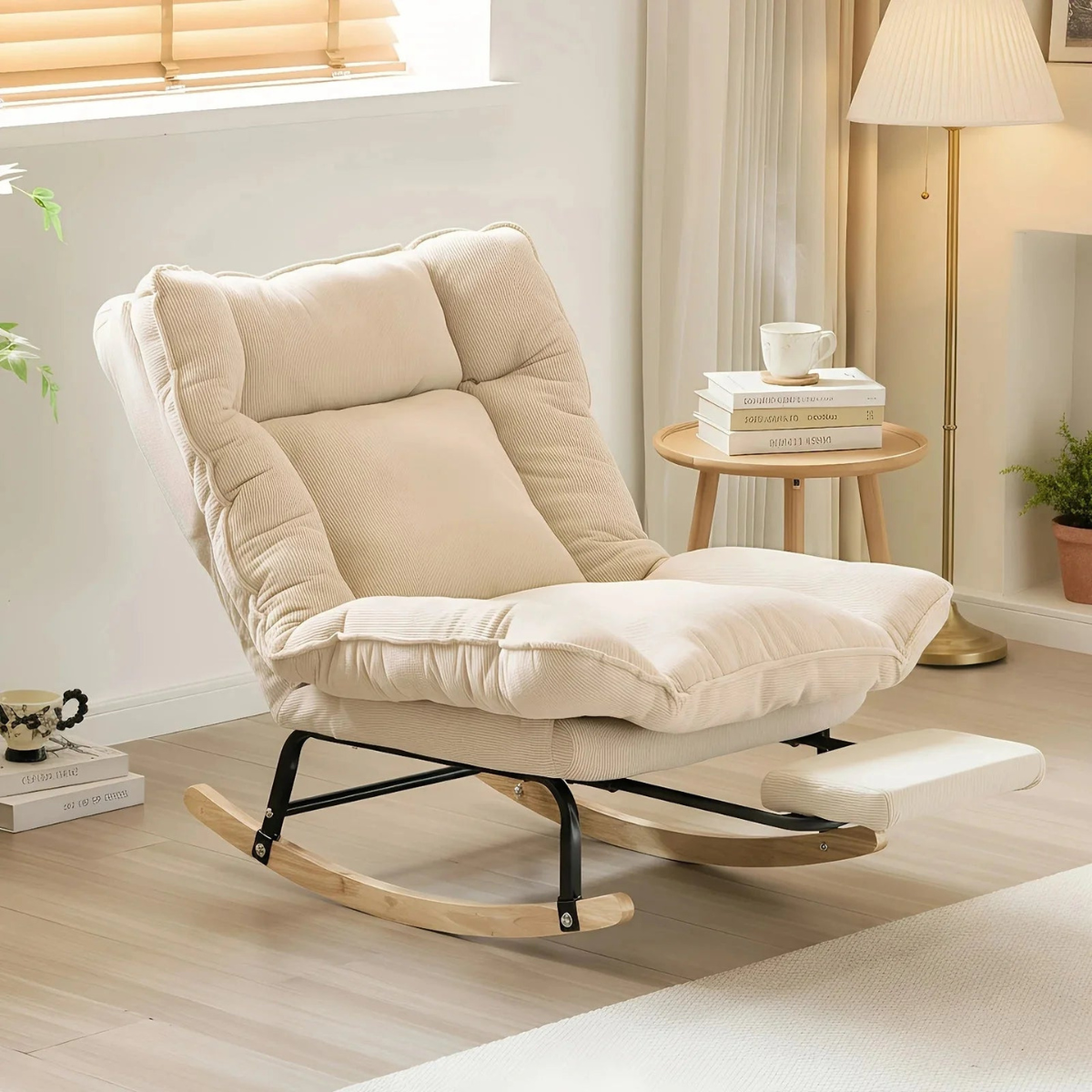 A cream-colored cushioned rocking chair with a footrest, set in a bright, modern living room next to a small wooden side table and floor lamp.