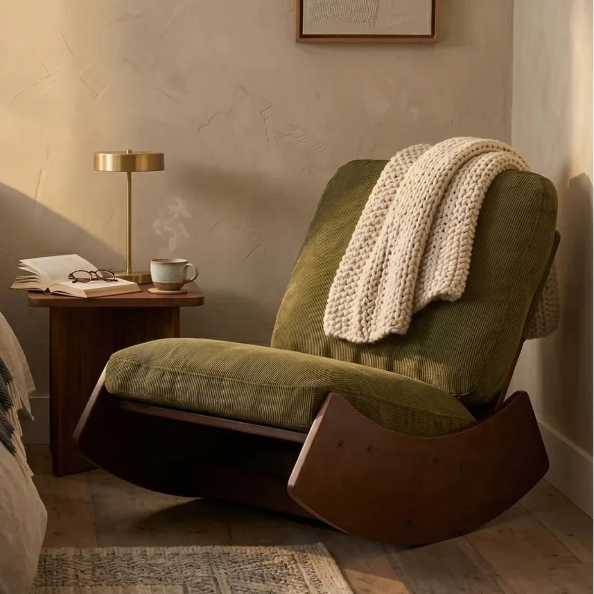 An olive green upholstered rocking chair with a dark wood base, styled in a warm room with a cream knit throw blanket draped over the back, a wooden side table, and a brass table lamp.