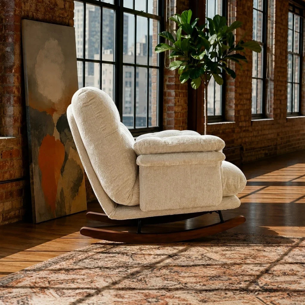A cream upholstered recliner chair with a matching ottoman and wooden rocking base, placed in a sunlit loft room with brick walls, large windows, a plant, and a patterned rug.