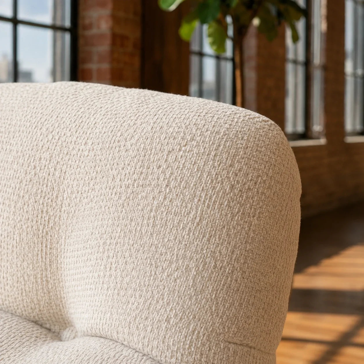 Close-up of a cream-colored textured bouclé armchair in a bright loft-style room with brick walls, large windows, and warm sunlight.