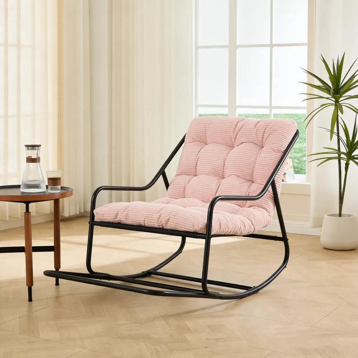 A modern rocking chair with a black metal frame and a tufted light pink striped cushion, set in a bright room with a side table and a potted plant.