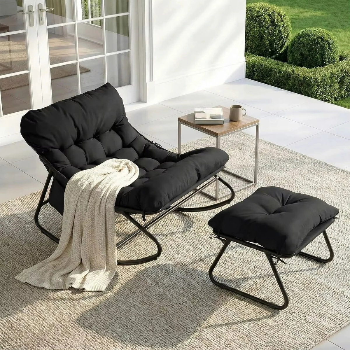 A black outdoor lounge chair with a matching ottoman and a small side table, styled on a textured rug in a bright patio setting with a white door and green hedges in the background.