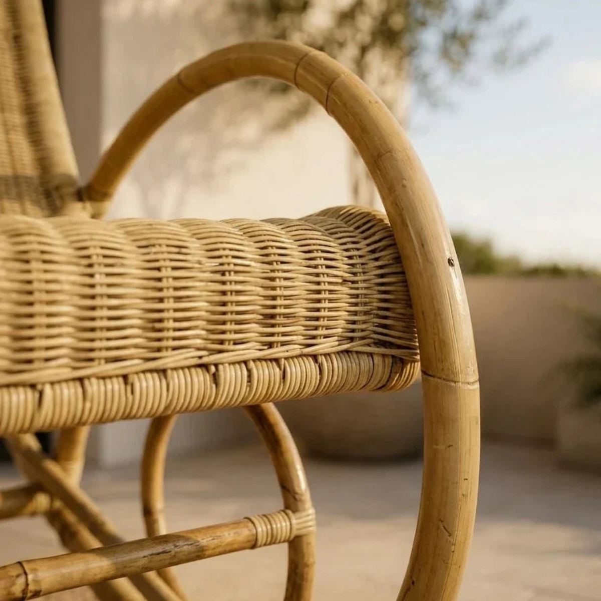 Close-up of a natural woven rattan chair with a curved bamboo armrest and seat, photographed outdoors with a softly blurred neutral background.