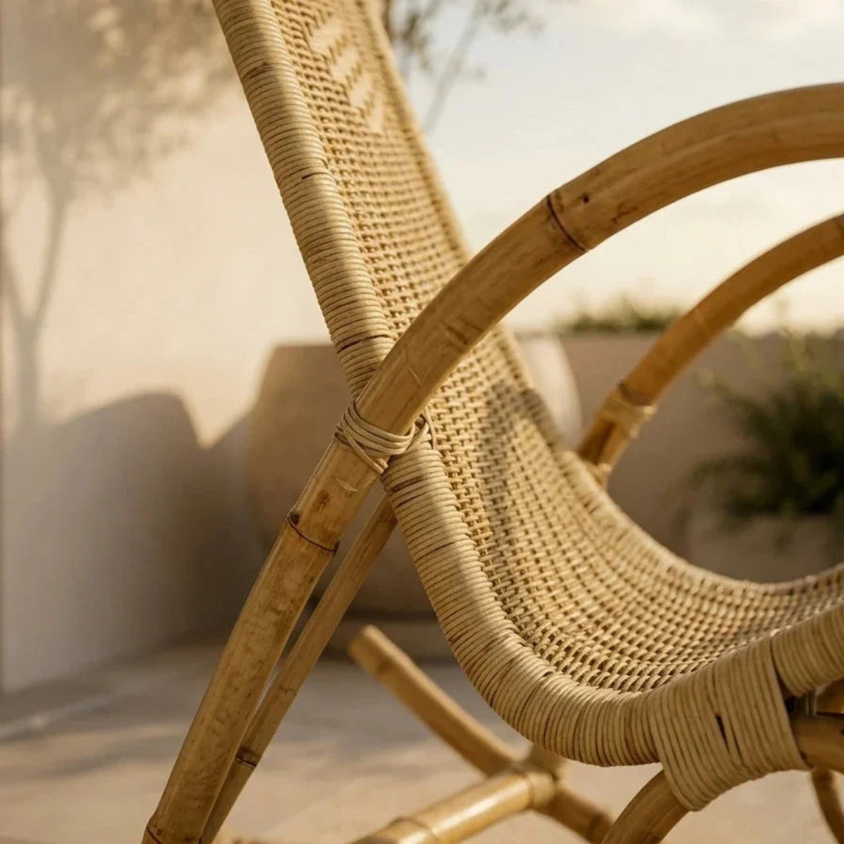 A close-up of a rattan or wicker rocking chair in a warm natural beige tone, photographed in a softly lit outdoor setting with blurred walls and plants in the background.