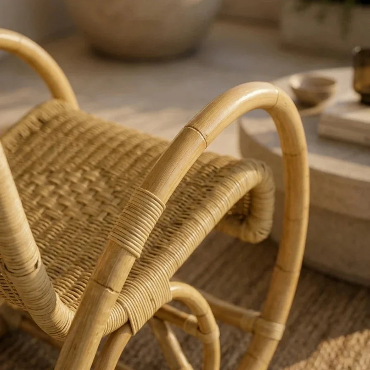 Close-up of a rattan or wicker chair with curved light tan wooden arms and a woven seat, photographed in a softly lit indoor setting.