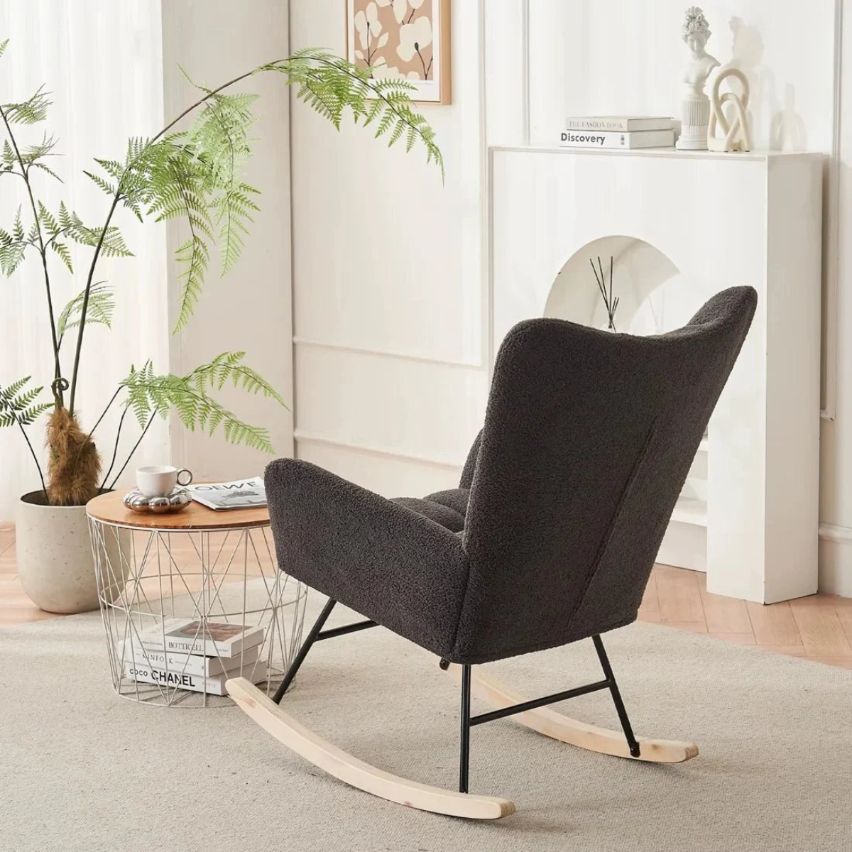 A dark charcoal gray upholstered rocking chair with light wood rockers and black metal legs, styled in a bright, modern living room next to a wire-frame side table.