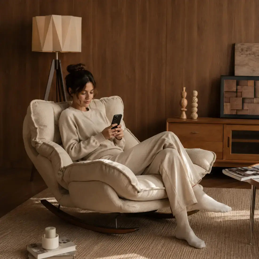 A woman in a matching beige lounge set sits in a plush cream-colored rocking chair in a warm, wood-paneled living room, holding a smartphone.