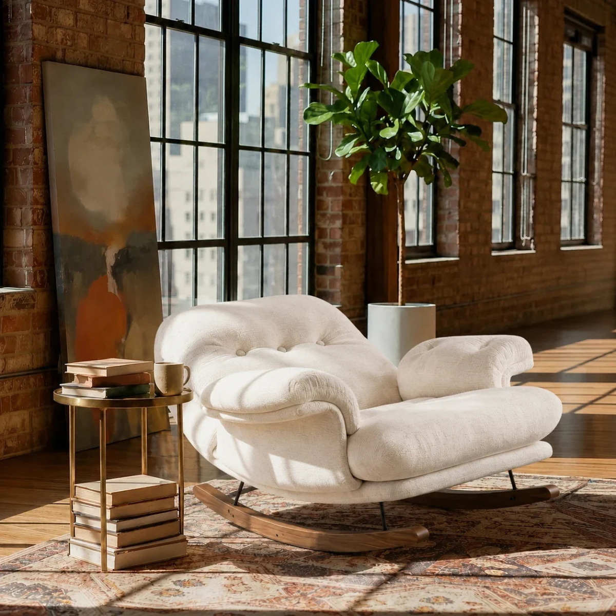 A cream-colored upholstered rocking chair with tufted backrest and wooden rocking bases, placed in a sunlit loft room with brick walls, large windows, a potted plant, and a small side table.