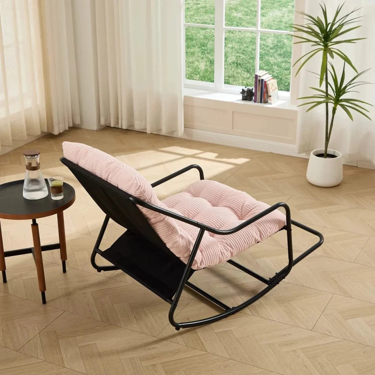 Black rocking chair with pink cushion on a wooden floor in a bright room with a window and plant.