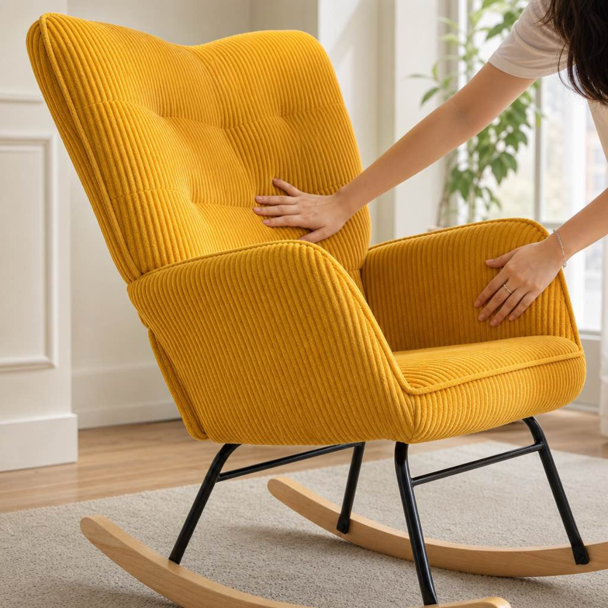 A mustard yellow corduroy rocking chair with a high tufted back, shown in a bright room with a woman's hands touching the fabric. The chair has black metal legs and a natural wood rocking base.