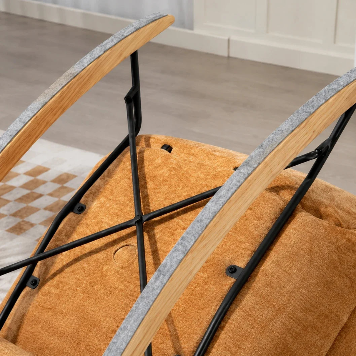 Close-up of a modern lounge chair showing the tan textured fabric seat, curved light wood armrests with gray felt edging, and a black metal frame in an indoor room setting.