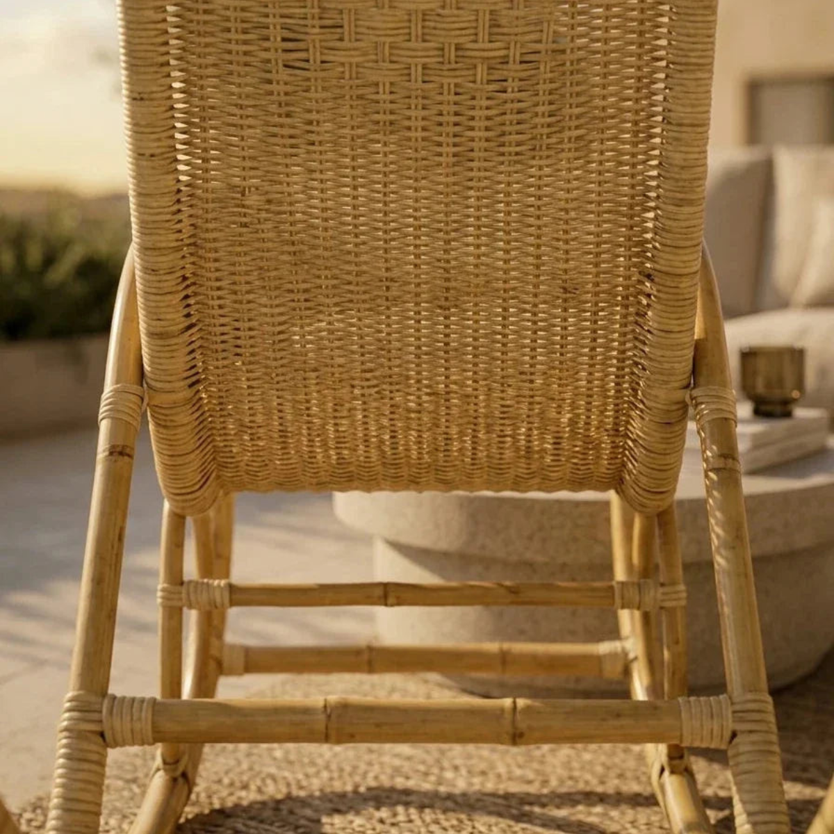 A close-up of a natural woven rattan or wicker chair back and frame, shown in a warm outdoor patio setting with a blurred sofa and table in the background.
