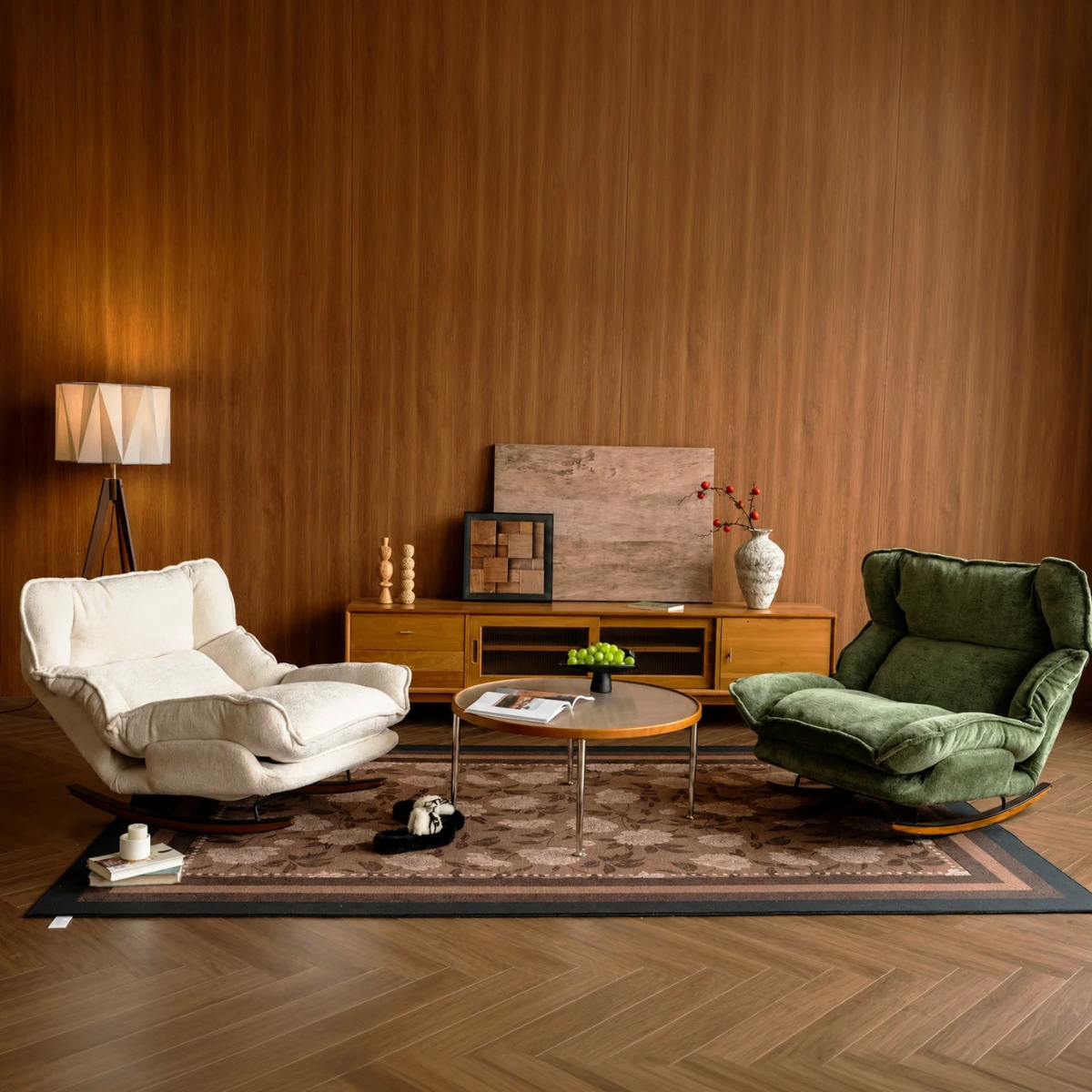 A styled living room scene with two upholstered rocking chairs facing a round coffee table, set on a patterned area rug against a wood-paneled wall. One chair is off-white and the other is deep green, with a wooden cabinet, lamp, and decor in between.